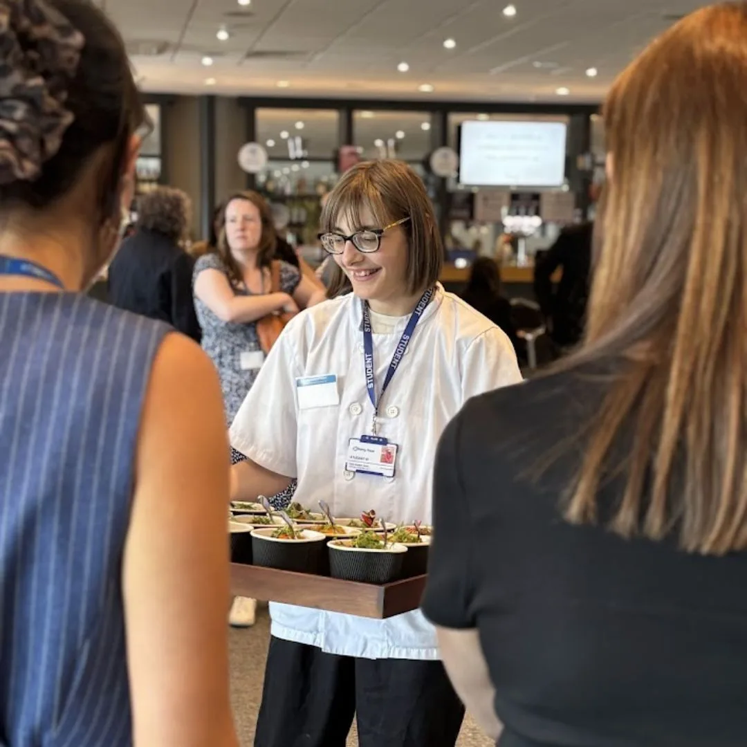 A server in a white chef's jacket and lanyard carrying a tray of food bowls through a crowded room of people wearing name badges